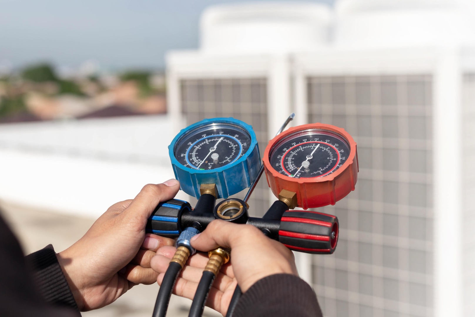 about-01 Person holding a manifold gauge set with blue and red dials, measuring pressure in HVAC systems—perfect for your landing page header, with outdoor equipment visible in the background.