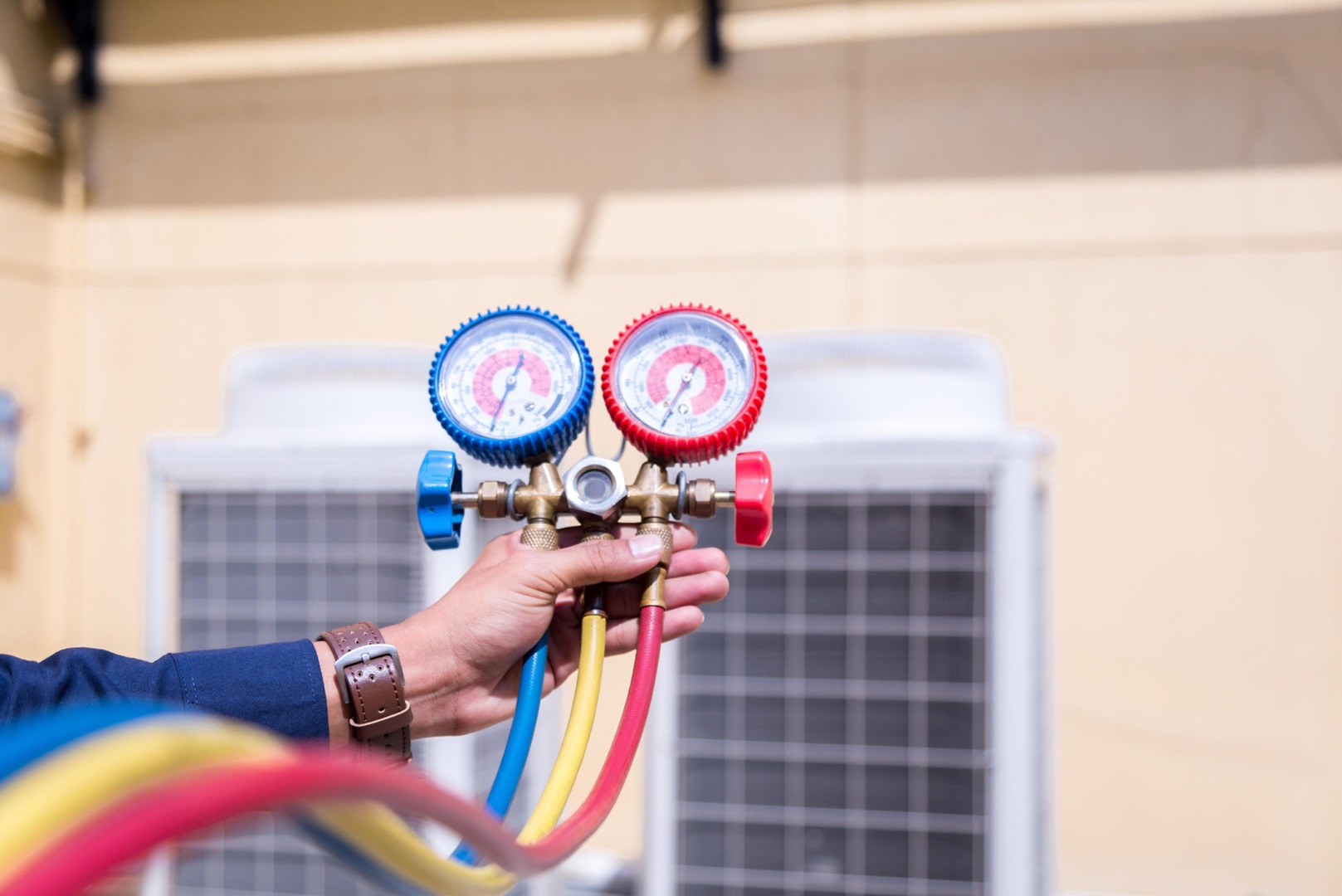 services-03 A person holds HVAC manifold gauges with red, blue, and yellow hoses, in front of an outdoor air conditioning unit—perfect for a landing page header image.