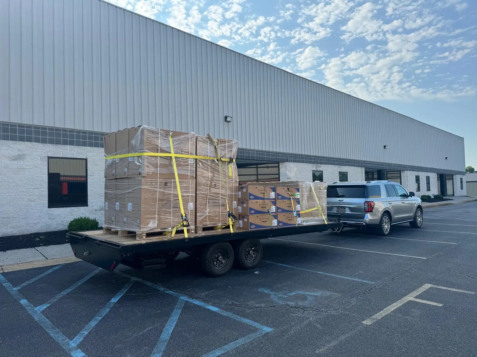 why-choose-us-02 A silver SUV, possibly belonging to an air conditioning repairman, tows a flatbed trailer loaded with large, shrink-wrapped pallets and boxes outside a warehouse building.