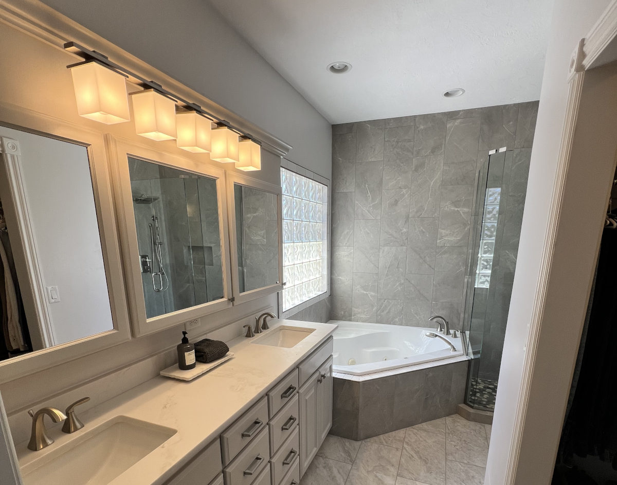 modern-bathroom-remodel-white-double-vanity-grey-tile A modern, spacious bathroom featuring a white double vanity with rectangular mirrors, warm square lighting fixtures, and a corner jetted bathtub surrounded by large grey marble-look tiles