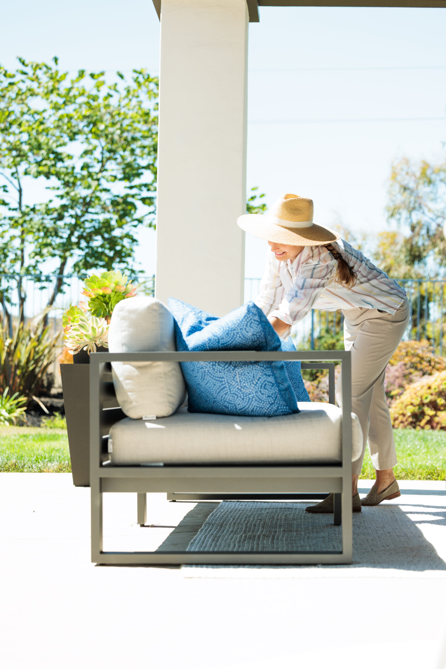Fixing up the outdoor living room Woman in a Ralph Lauren straw hat fluffing blue and beige pillows on a modern aluminum outdoor couch with green trees in the background.