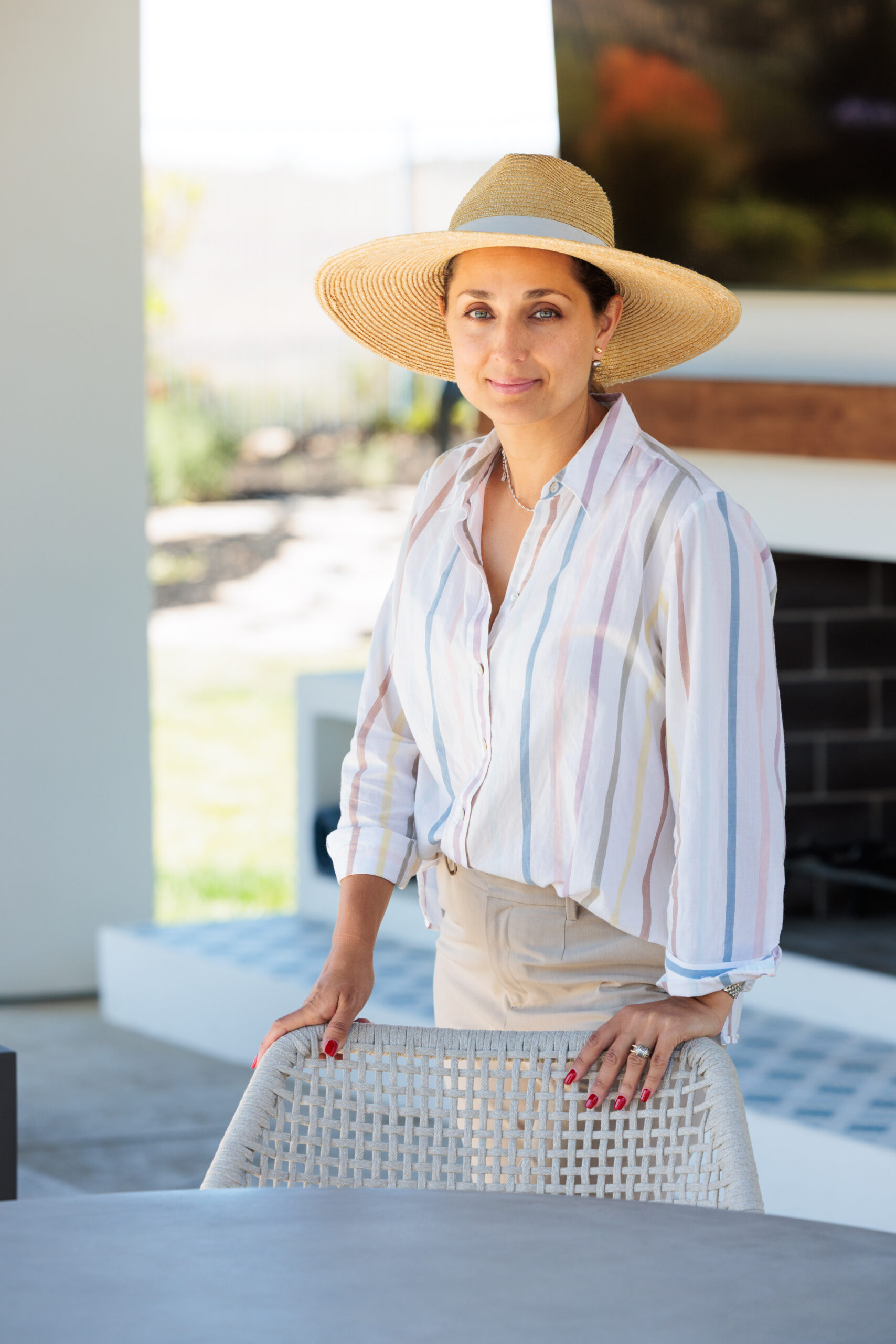 Kristina Jones Kristina Jones standing by an outdoor dining table and holding onto a chair, wearing a Ralph Lauren straw hat, light stripped cotton button-down shirt and beige pants. Outdoor fireplace with white stucco in the background with TV hanging on the fireplace and wood mantle under the TV.