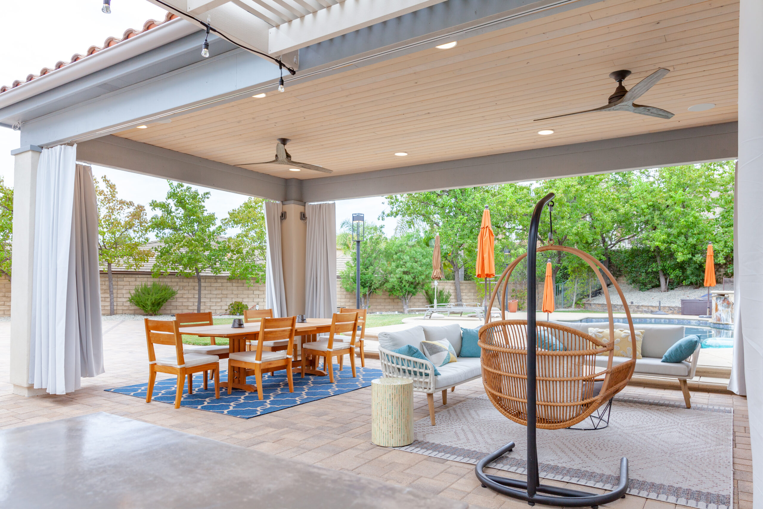 Custom Poolside Cabana with Automated Curtains | Newbury Park, CA Luxury outdoor cabana in Newbury Park with wood-panels ceiling, ceiling fans, recessed lighting, automated curtains, outdoor dining area, lounge seating, and pool views.