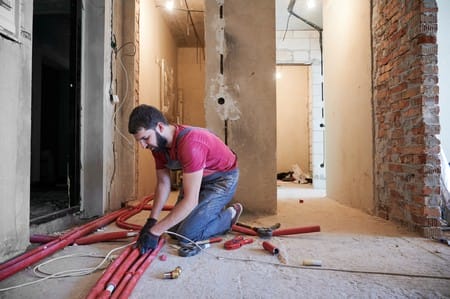 How Much Does Plumbing Cost for a New House? 2 Young bearded plumber installing underfloor heating system in new apartment