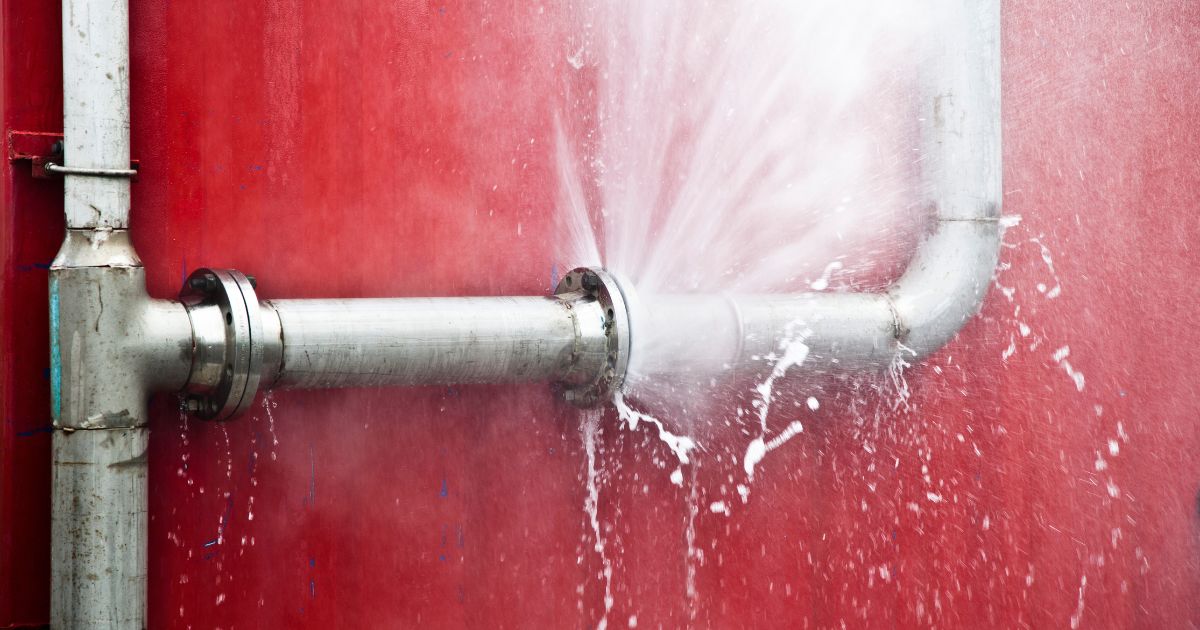 A metal pipe joint spraying a high-pressure stream of water against a bright red wall, indicating a burst pipe.