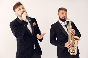 about-02 Two male musicians in black suits performing with a saxophone and microphone on a studio background.
