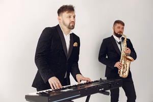 hero-02 Two male musicians in suits playing a keyboard and saxophone in a studio setting.