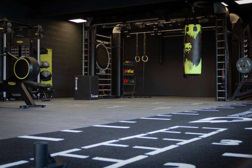 Une salle de sport bien รฉquipรฉe avec des รฉquipements d'exercice noirs et jaunes, notamment un sac de boxe, des anneaux, des poids et plusieurs stations de fitness sur un sol dรฉlimitรฉ.
