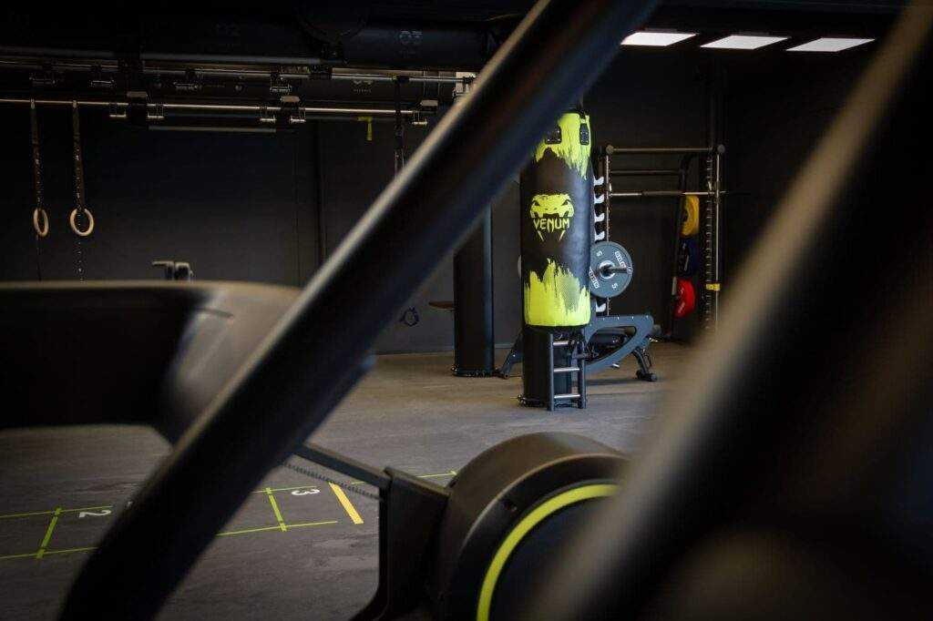 Une salle de sport avec un sac de boxe, un tapis roulant, du matรฉriel d'haltรฉrophilie et des anneaux de gymnastique. La piรจce est faiblement รฉclairรฉe avec un design moderne et industriel.