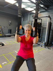 Gema, vรชtue d'une chemise rouge, sourit ร la camรฉra tout en utilisant des sangles de suspension TRX dans une salle de sport.