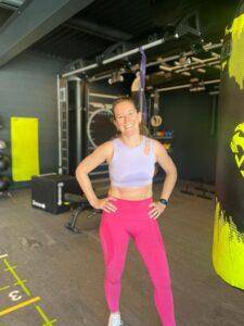 Isa, une femme vรชtue d'un soutien-gorge de sport violet et de leggings roses, sourit ร la camรฉra dans une salle de sport. L'รฉquipement d'exercice est visible en arriรจre-plan.