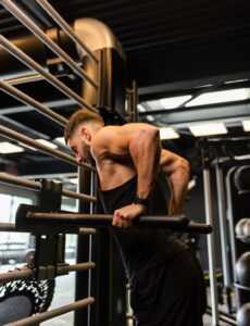 Une personne dans une salle de sport effectue un plongeon des triceps sur des barres parallèles, vêtue d'un débardeur et d'un short noirs, avec un équipement de gymnastique visible en arrière-plan.
