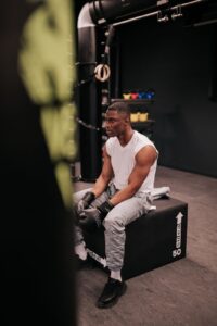 Un homme vêtu d'une chemise sans manches blanche et d'un pantalon gris est assis sur une boîte noire, portant des gants de boxe noirs, dans une salle de sport. Les équipements de fitness sont visibles en arrière-plan.
