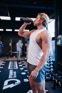 Un homme dans une salle de sport boit avec une bouteille d’eau. Il porte une chemise blanche sans manches et un short gris, et il y a du matériel de gym en arrière-plan.