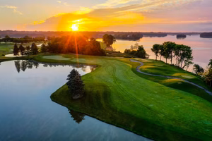 A sunrise over a tranquil golf resort in Ypsilanti, MI, illuminates the lush green fairways and shimmering pond, framed by trees and a distant shoreline.