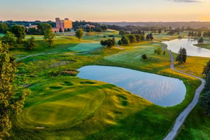 Aerial view of a golf course in Ypsilanti, MI, showcasing lush green fairways, a sand trap, and a serene pond. Trees dot the landscape, with a building visible in the distance under the clear sky, offering the perfect setting for both outdoor play and golf simulator practice.