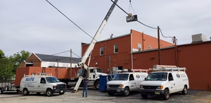 A crane lifts equipment onto the roof of a red brick building, possibly installing a Building Automation System, as three white HVAC service vans are parked nearby and a worker directs the operation.
