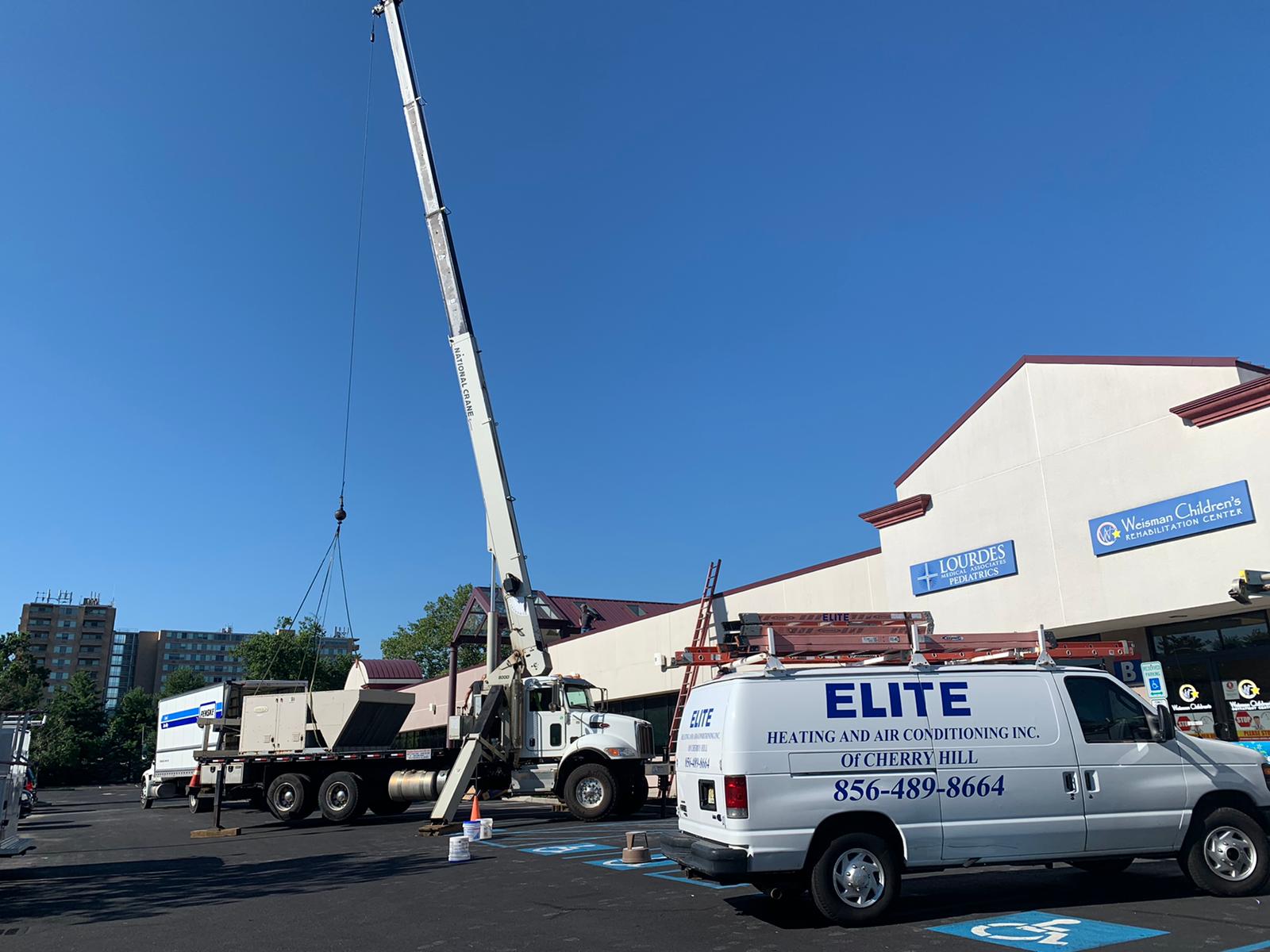 A crane lifts equipment beside Elite Heating and Air Conditioning vans parked outside a commercial building, preparing for the installation of a Building Automation System under clear skies.