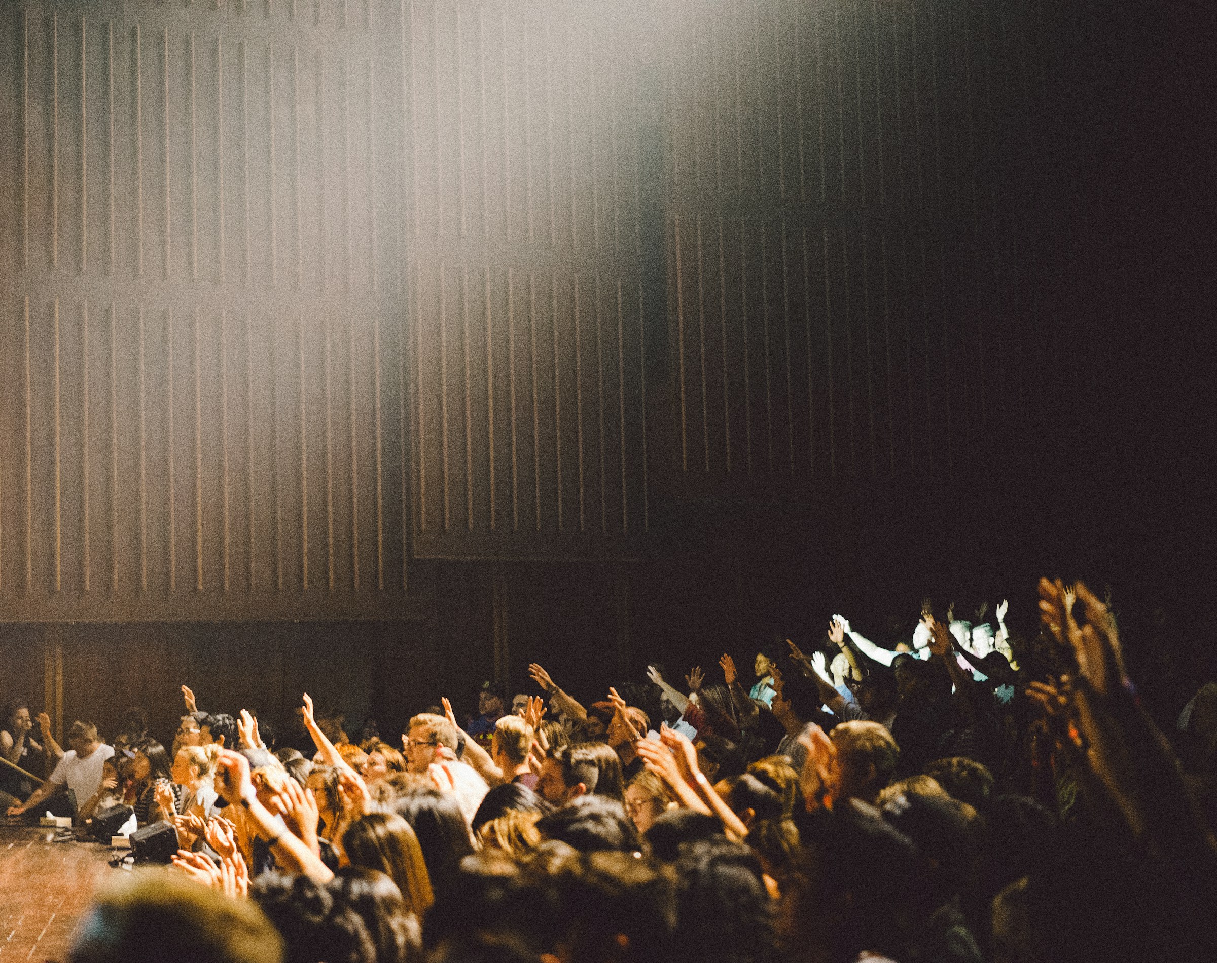 Audience members seated indoors raise their hands toward a brightly lit stage, surrounded by dark wooden panels and enhanced comfort thanks to a modern Building Automation System.