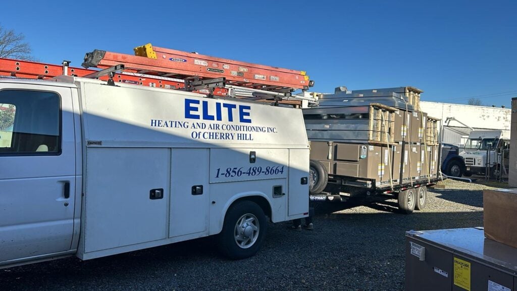 A white ELITE Heating and Air Conditioning van with ladders on top is parked next to a trailer loaded with HVAC units in a gravel lot, ready for Building Automation System installation.
