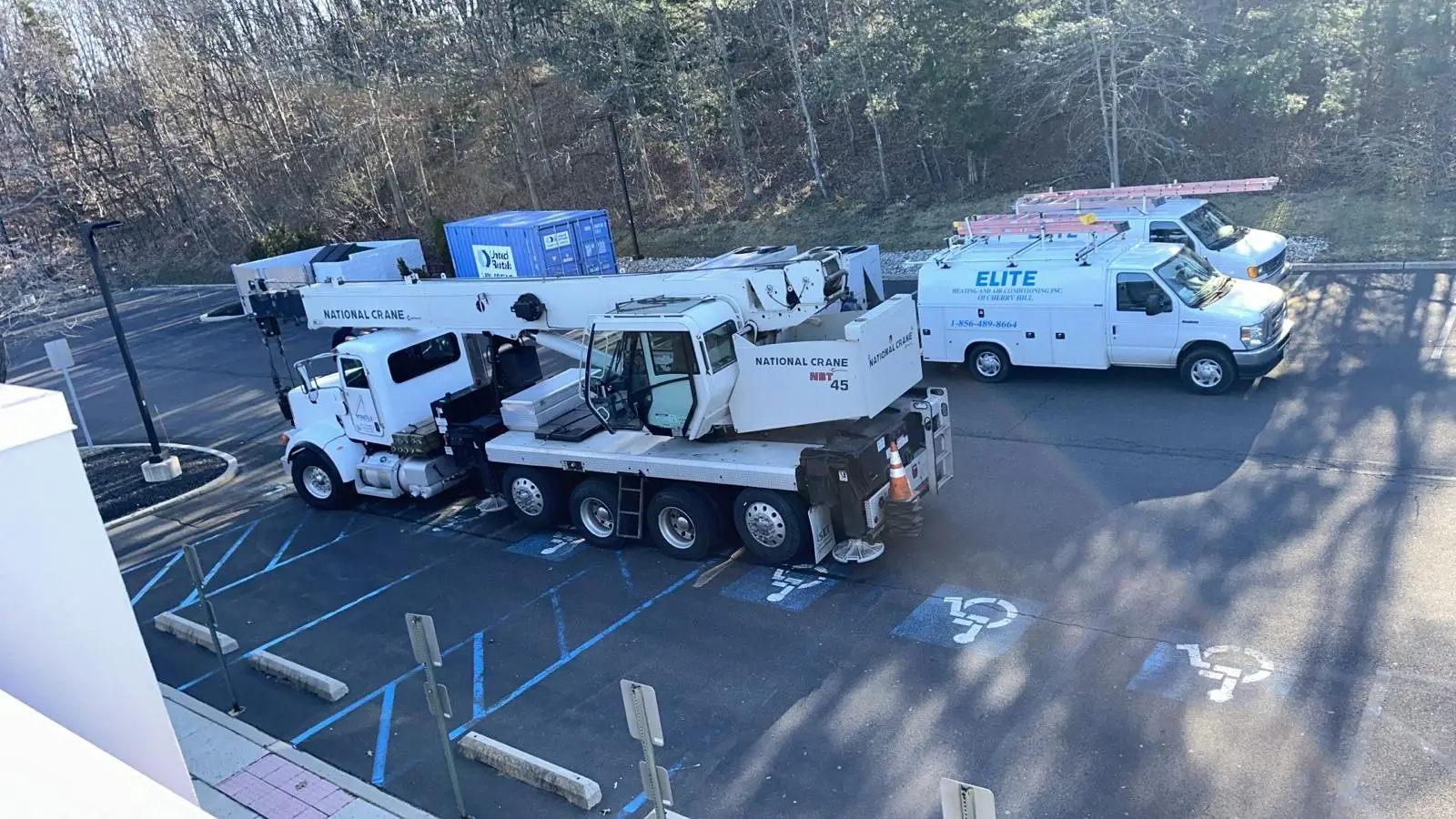A National Crane truck and an Elite Electrical van, both used for commercial AC repairs, are parked across multiple handicap spaces in a mostly empty lot near trees.