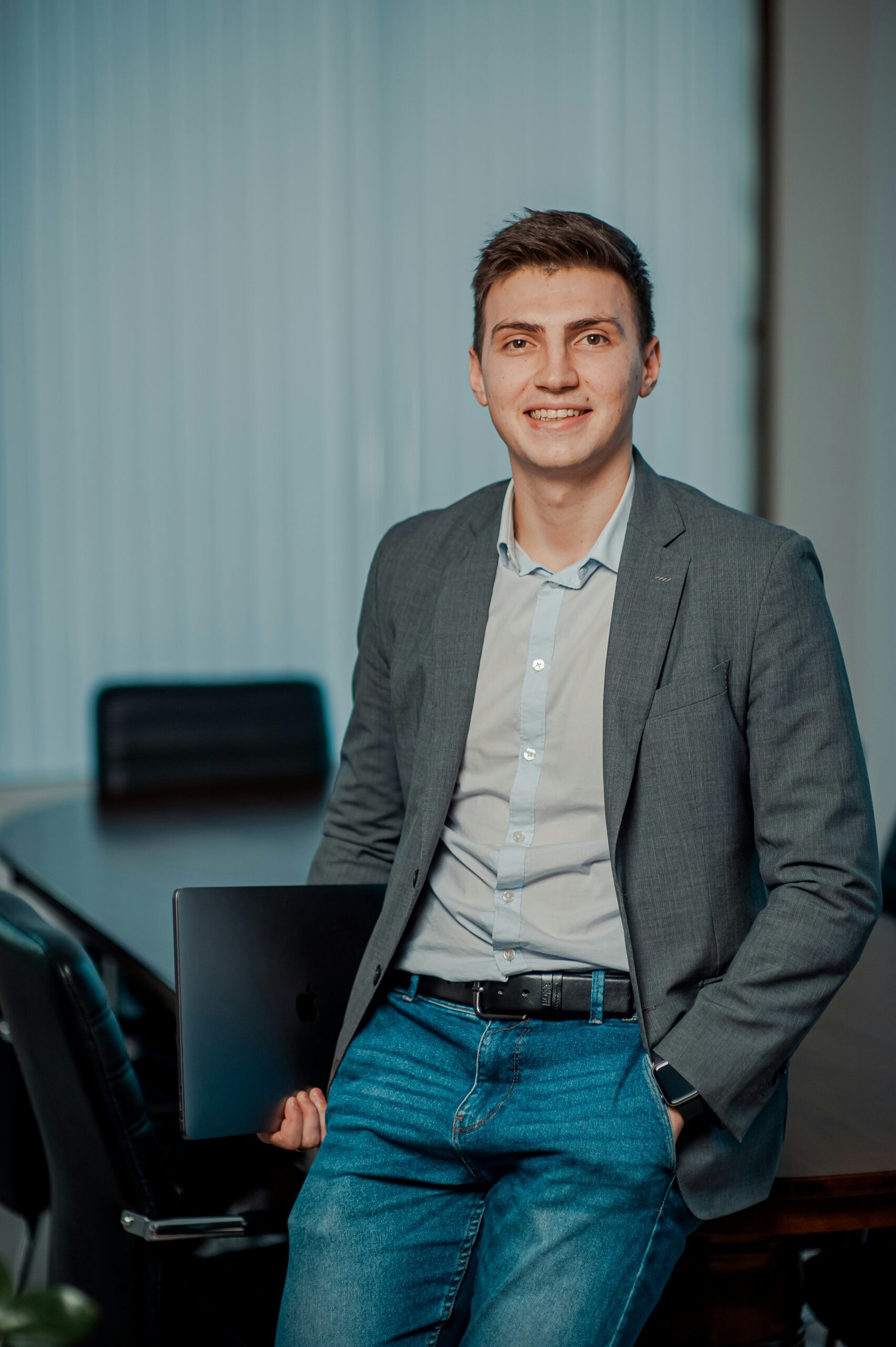 A man in a gray blazer and jeans stands in an office, holding a closed laptop and smiling at the camera—capturing one of those unusual moments in a busy commercial HVAC firm.