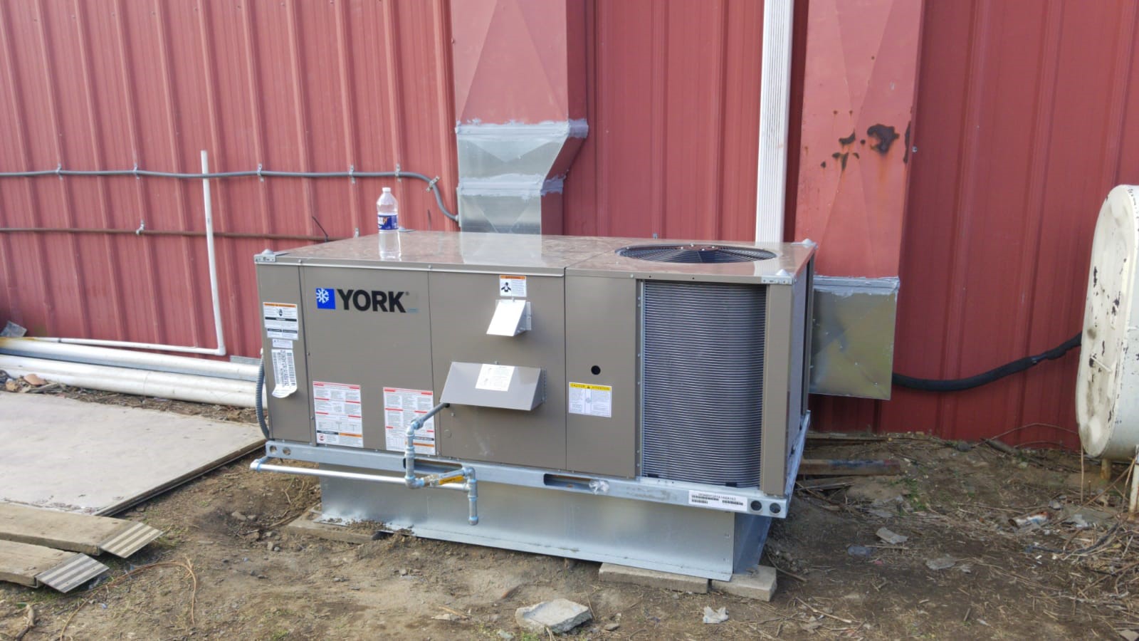 A YORK Commercial HVAC unit is installed on a metal platform outside a red corrugated metal building; pipes and wiring are visible along the wall, capturing an unusual moment in 2025 industrial design.