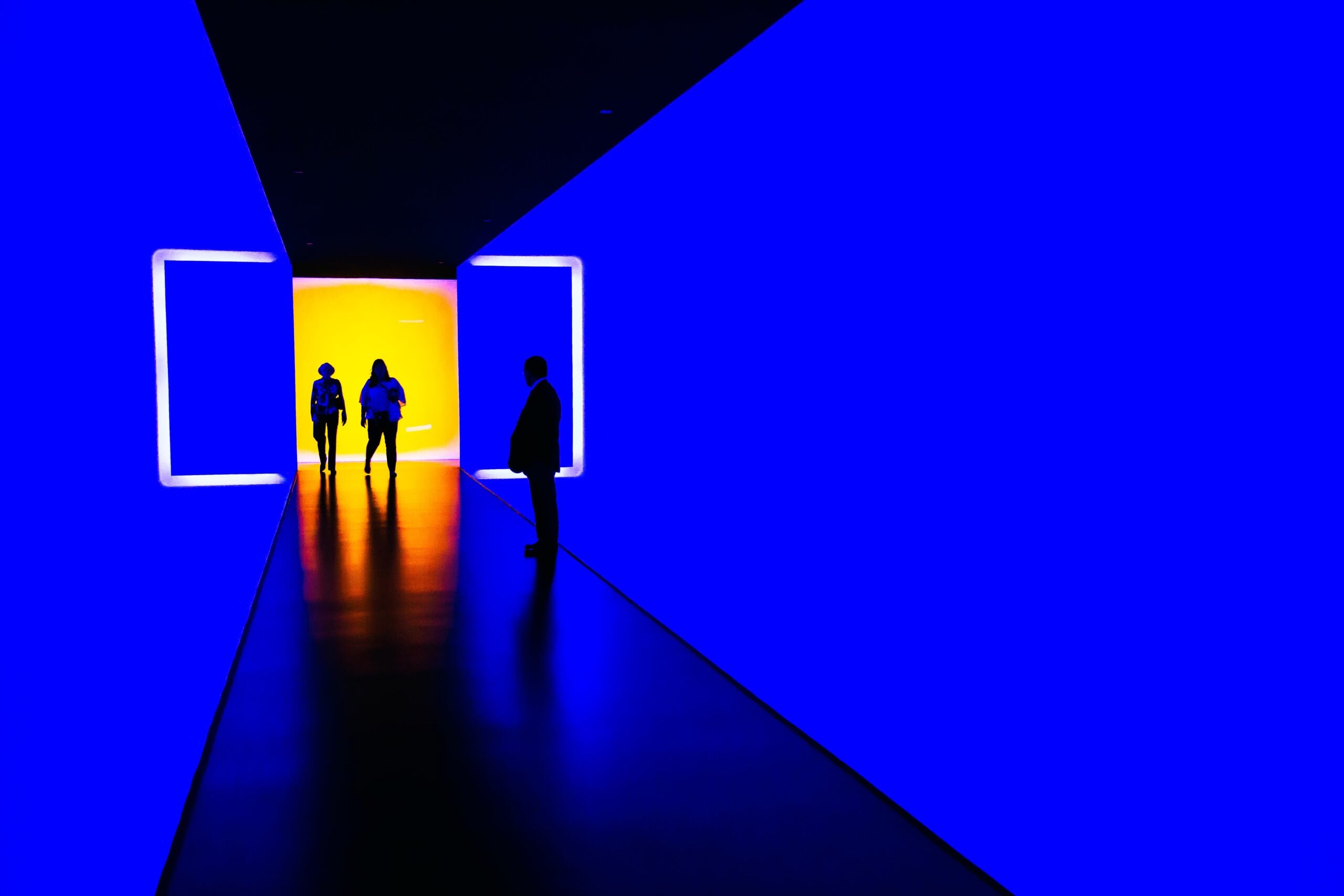 Silhouetted figures walk through a hallway with bright blue and yellow illuminated walls, creating a stark contrast between light and shadow—an unusual moment in the world of commercial HVAC design for 2025.