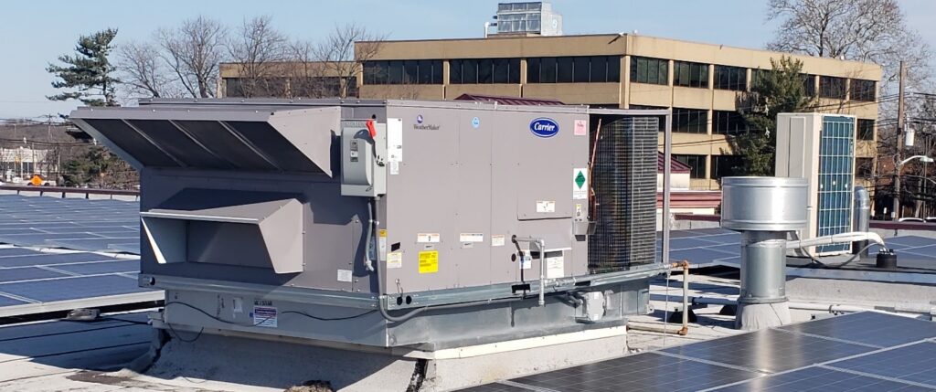 A large commercial HVAC unit sits next to solar panels and other equipment atop a rooftop, with office buildings and trees in the background—capturing one of 2025’s unusual moments in sustainable design.