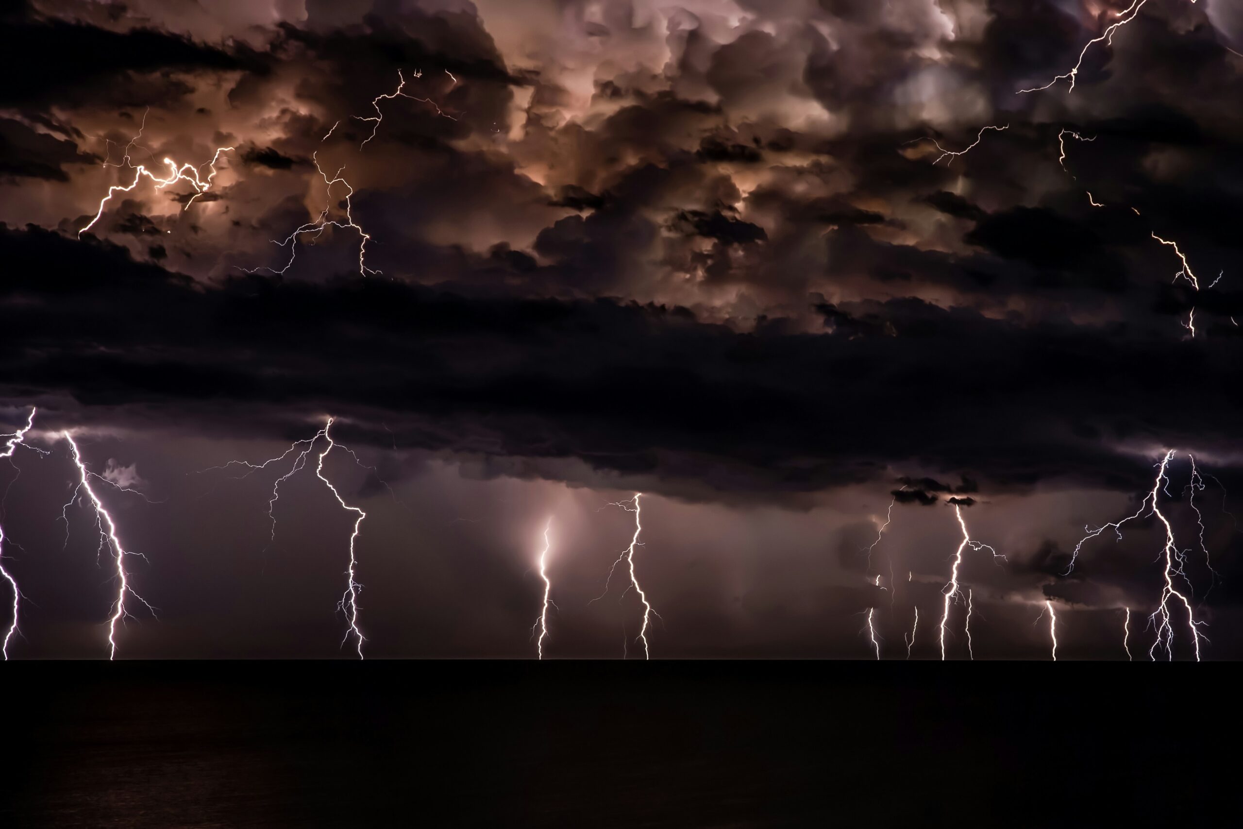 Multiple lightning bolts illuminate dark storm clouds over a calm body of water at night.