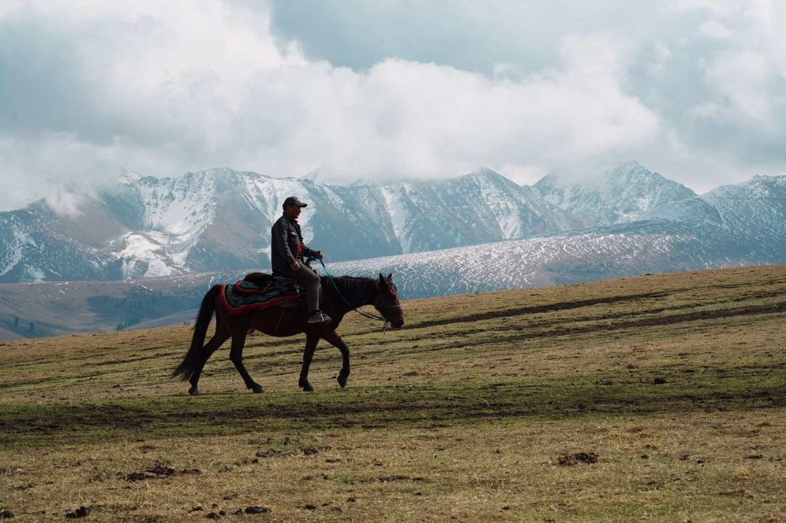 A rider in the saddle crossing an open fell slope with a long snow-capped range behind