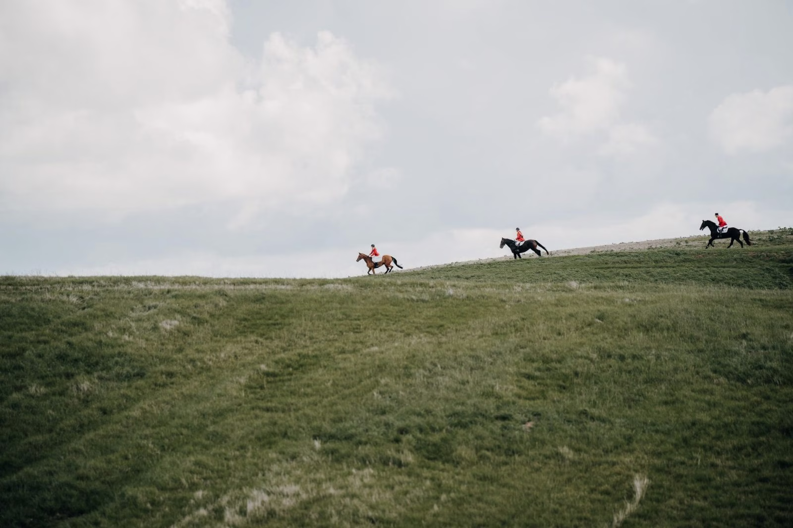Three riders spread along the skyline of an open grass ridge beneath a wide cloudy sky