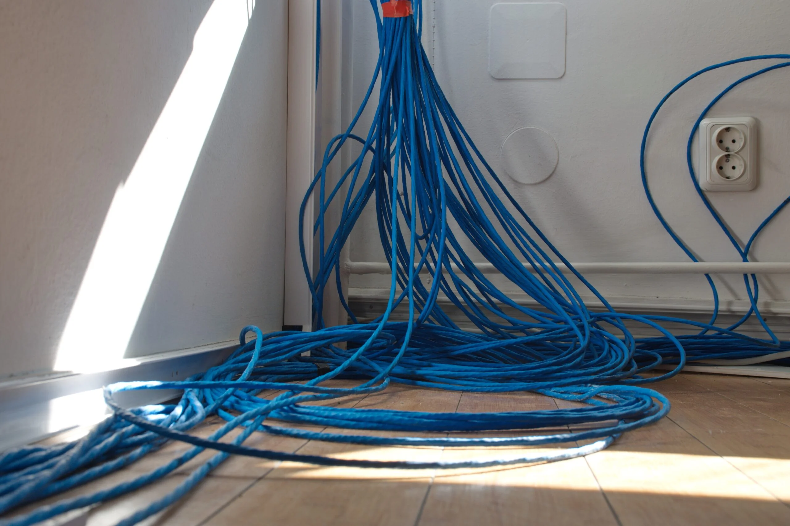 Numerous blue network cables are gathered in the corner of a room, trailing along the floor near a power outlet and white walls—a scene fit for any professional guide on organizing tech spaces in Scottsdale.