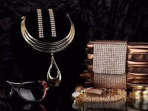 Une présentation de bijoux et d'accessoires en or sur un fond en tissu noir, comprenant un collier tendance, des boucles d'oreilles assorties, une pochette en or, des lunettes de soleil et des bracelets - Ce que j'achète quand on s'offre du luxe.