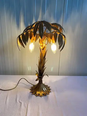 Une lampe de table décorative en forme de palmier, avec une base et des feuilles dorées, est exactement ce que j'achète. Elle est dotée de trois ampoules lumineuses suspendues sous le feuillage et repose sur une surface blanche contre un mur lambrissé.