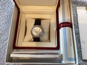 Une montre de luxe au bracelet foncé est présentée dans un coffret ouvert, accompagnée de livrets et d'un certificat. L'intérieur couleur crème met en valeur le nom de la marque « ZENITH » inscrit sur le certificat : un véritable symbole d'élégance et de sophistication.