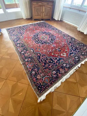 Un tapis rouge et bleu marine aux motifs riches et complexes est posé sur un parquet en bois dans une pièce où de grandes fenêtres, des rideaux blancs et une armoire en bois contre le mur créent une atmosphère chaleureuse. Idéal pour ceux qui recherchent une touche d'élégance.
