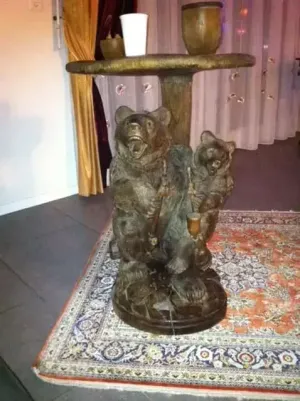 Une table en bois avec un support sculpté représentant deux ours, un grand et un petit, tenant des cannes à pêche. La table est élégamment dressée sur un tapis à motifs, avec une tasse blanche et un bol en bois, parfaits pour présenter vos achats en ligne.