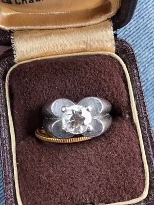 Une bague en argent ornée d'un gros diamant rond au design incurvé unique est présentée dans un écrin marron doublé de velours. Le couvercle intérieur couleur crème contraste avec l'extérieur sombre, laissant clairement entendre : « Ce que j'achète. ».