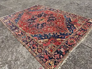 Un tapis persan coloré aux motifs complexes, rappelant « Ce que j'achète », repose sur une surface en béton brut. Il arbore des motifs géométriques aux riches nuances de rouge, bleu, jaune et crème, avec un médaillon central proéminent et des bordures ornées.