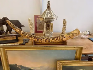 Une défense en ivoire finement sculptée est exposée sur un support en bois. Derrière elle, un objet décoratif détaillé et une figurine d'éléphant, rappelant les objets ménagers rares, sont exposés. Plusieurs œuvres d'art encadrées sont partiellement visibles au premier plan, contribuant à l'ambiance soignée.