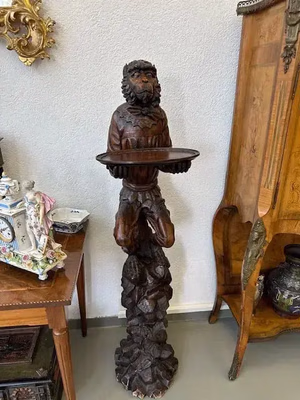 Une sculpture en bois représentant un personnage debout tenant un plateau est placée dans un coin de la pièce. Cette sculpture complexe, représentant « ce que j'achète », présente des textures détaillées et est entourée de meubles et d'objets décoratifs.