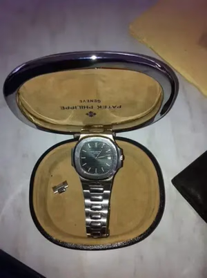 Une montre-bracelet de luxe avec un bracelet en métal argenté est présentée dans un boîtier ovale marron au pourtour argenté brillant. L'intérieur du boîtier porte l'inscription « Patek Philippe Genève ». Ce que j'achète comprend également un petit outil à côté de la montre.
