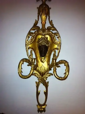 Une applique murale dorée ornée de motifs complexes en forme de feuilles et de deux bougeoirs incurvés est exactement ce que j'achète. En son centre, un petit visage sculpté aux tons sombres porte une coiffe décorative, élégamment placé sur un mur blanc immaculé.