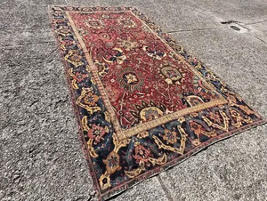 Un grand tapis persan orné avec un riche fond rouge présentant des motifs floraux complexes en or, bleu marine et beige - vraiment représentatif de ce que j'achète - est magnifiquement présenté sur une surface en béton texturé.