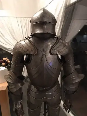 Une armure médiévale est exposée à l'intérieur, composée de plaques métalliques sombres, d'un casque à visière fermée et d'articulations. Elle ne ressemble à aucun autre objet ordinaire : un véritable chef-d'œuvre qui s'intègre parfaitement à la pièce tamisée et aux rideaux transparents.