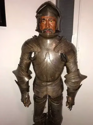 Dans une pièce, un mannequin grandeur nature porte une armure ancienne en métal sombre, finement détaillée et ornée d'un casque à visage humain. Placée contre un mur clair, elle est idéale pour ceux qui recherchent le summum de la décoration historique.