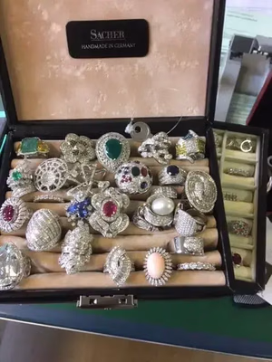 Un coffret à bijoux s'ouvre pour révéler diverses bagues, chacune ornée de pierres précieuses telles que diamants, émeraudes, rubis et perles. Ces pièces aux designs raffinés sont présentées dans un écrin doublé de velours estampillé « Sacher Handmade in Germany ». Idéal pour ceux qui privilégient l'élégance artisanale locale.
