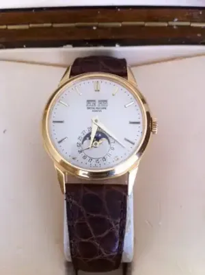 Cette montre-bracelet en or, dotée d'un cadran blanc orné de l'inscription « Patek Philippe » en haut, d'un indicateur de date automatique avec sous-cadran et d'un bracelet en cuir marron, repose élégamment sur une surface beige. Ce garde-temps exquis incarne le luxe et un design intemporel.
