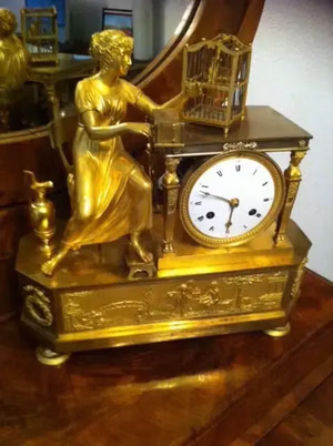 Cette horloge ancienne en or, richement détaillée et dotée d'un cadran à chiffres romains, représente une femme sculptée en robe fluide tenant une cage à oiseaux. Des motifs et des colonnes ornent la base de cette pièce exquise, digne d'un achat par tout collectionneur. L'horloge est élégamment posée sur un plateau en bois.